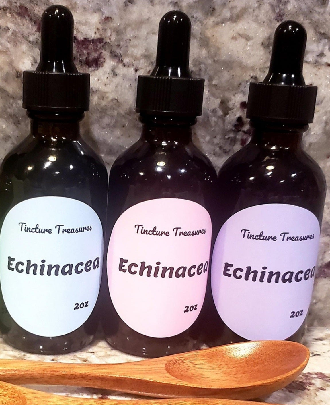 Three dark amber glass dropper bottles labeled 'Tincture Treasures Echinacea 2oz' with black droppers, arranged on a stone-like surface with two wooden spoons in front.