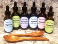 Six amber glass dropper bottles of Tincture Treasures Hyssop tinctures with black droppers and pastel labels, arranged on a marble surface with two wooden spoons in front.
