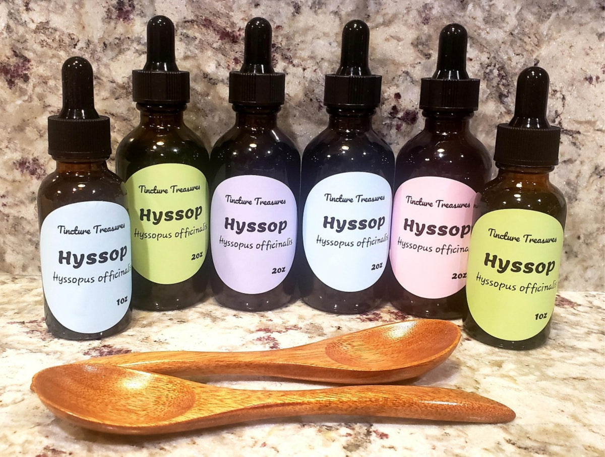 Six amber glass dropper bottles of Tincture Treasures Hyssop tinctures with black droppers and pastel labels, arranged on a marble surface with two wooden spoons in front.