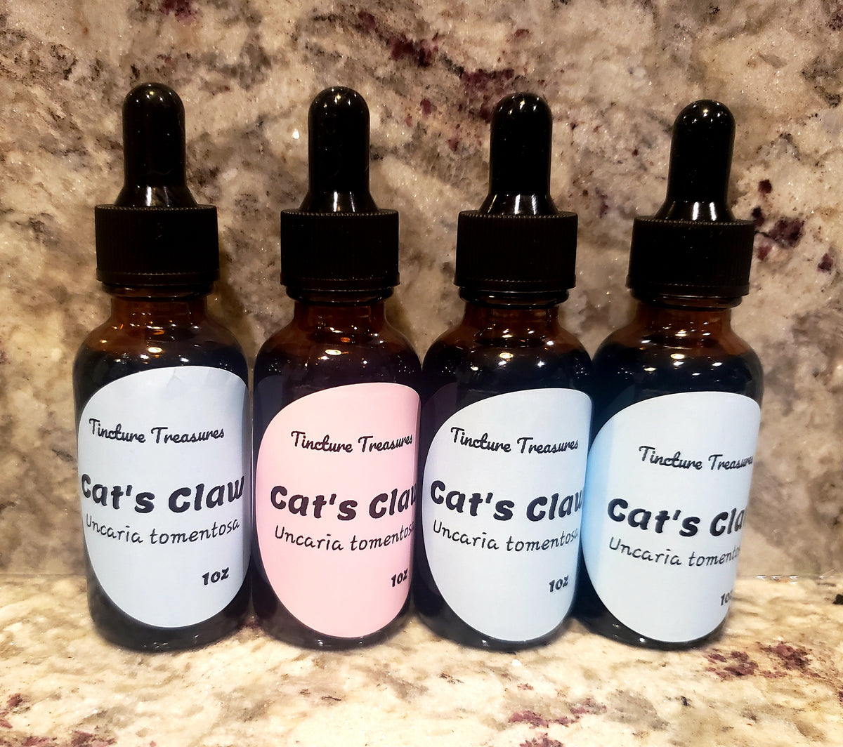 Four amber glass dropper bottles labeled 'Tincture Treasures Cat's Claw' arranged on a granite surface. Three bottles have blue labels and one has a pink label.