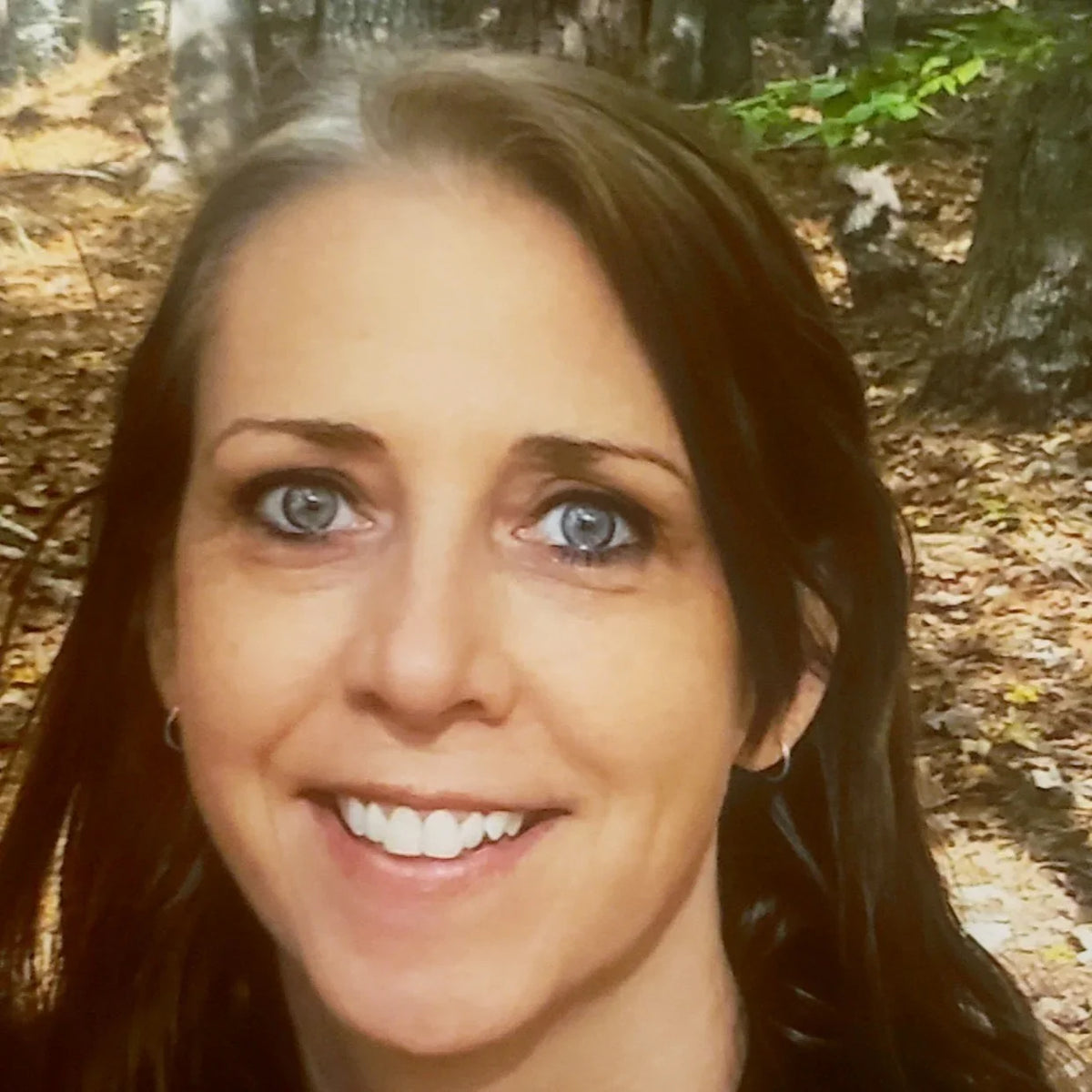 A close-up portrait of a smiling woman with long dark brown hair and blue eyes, photographed outdoors in a wooded setting with trees and leaf-covered ground in the background.